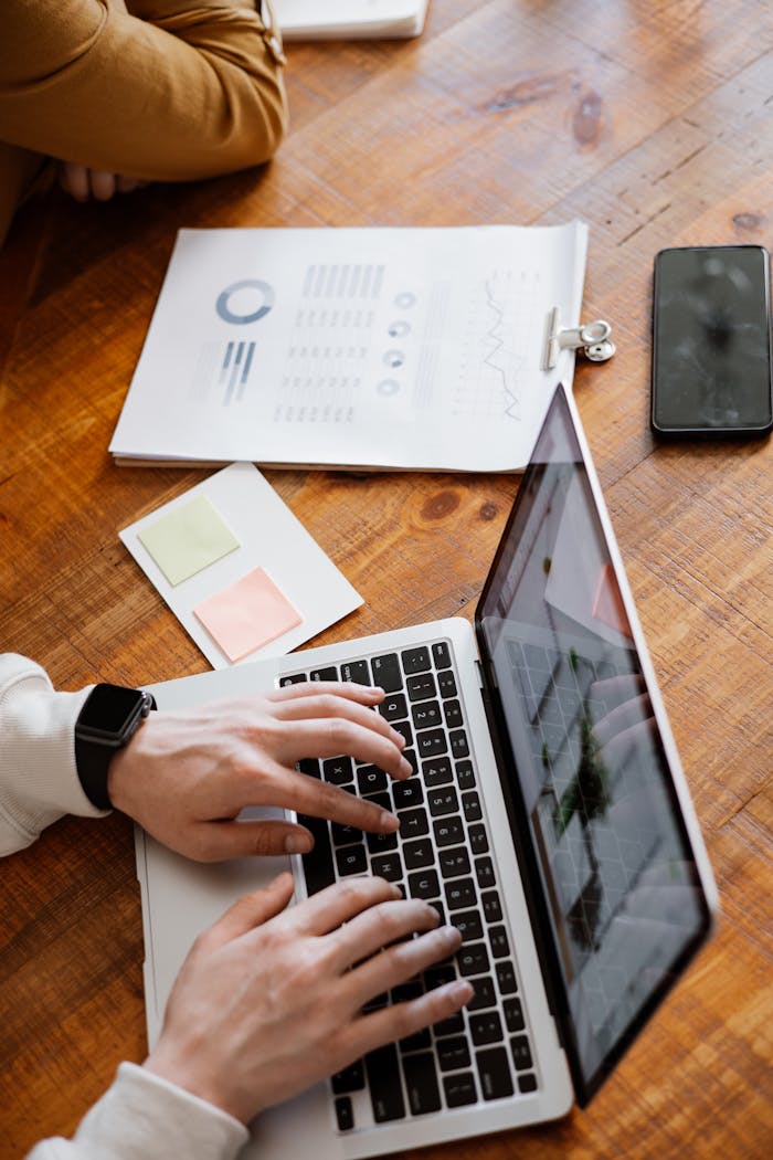 Hands typing on a laptop with paperwork and smartphone on a wooden desk, ideal for business and office themes.